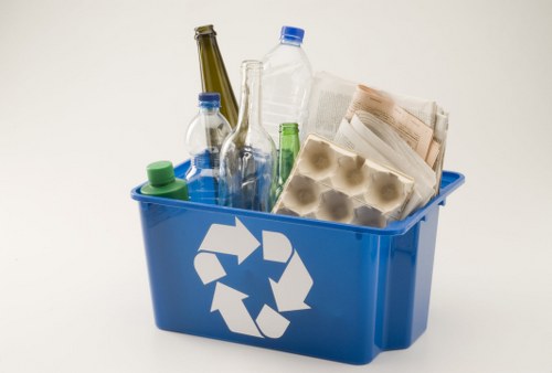 A blue plastic recycling bin with a white recycling symbol on its side, placed against a plain white background. The bin contains various recyclable waste items, including several transparent plastic bottles with blue, green, and clear caps, a carton of eggs with an egg-shaped compartment visible, and some folded newspapers or paper sheets. The bottles are of different shapes and sizes, with some showing residue marks and ridges, and are positioned upright within the bin. The newspapers appear slightly crumpled, lying on top of the bottles. The overall arrangement suggests a collection of recyclable household waste, typical of waste removal services in Ascot and surrounding areas. The lighting is even, highlighting the different textures and transparency of the plastic bottles and the glossy finish of the paper and egg carton, emphasizing the practicality of recycling in home or property clearance contexts.