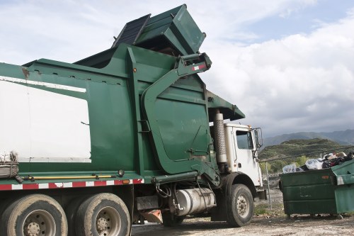 A large green skip loader truck is positioned on a gravel surface near a white building, with its hydraulic arm raised to unload a green skip. The skip has a rectangular shape with open top, made of metal, featuring a textured surface and visible weld lines. The truck's cab is white with a silver exhaust pipe, and the wheels are large with black tires, some of which show dirt and wear. In the background, a green open-top waste container filled with mixed rubbish is visible, situated outdoors with rolling hills, cloudy sky, and distant landscape suggesting a rural or semi-rural area possibly near Ascot or its surroundings. The overall scene reflects a waste collection process, typical of rubbish removal services such as those offered by House Clearance Ascot, with natural light illuminating the scene and emphasizing the textures of the metal and gravel.