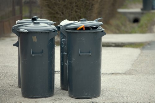 Two dark grey plastic wheelie bins with closed and open lids are positioned on a concrete driveway or pavement area. The bin on the left has a smooth surface and a visible handle, while the bin on the right has its lid partially open, revealing an orange-handled tool or object inside. Behind the bins, there is a background of blurred greenery and a section of a concrete bridge or overpass, indicating an outdoor residential or semi-urban environment. The scene appears to be part of waste removal or rubbish collection, consistent with services offered by House Clearance Ascot. The lighting is natural, suggesting daytime, with no significant shadows or reflections. The overall setting is tidy, and the bins are placed in an accessible position for rubbish disposal or collection.