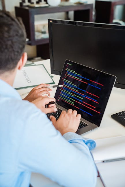 A person working at a desk with a laptop displaying a code editor filled with colorful lines of programming code. The individual, dressed in a light blue shirt, uses a mouse with their right hand whil