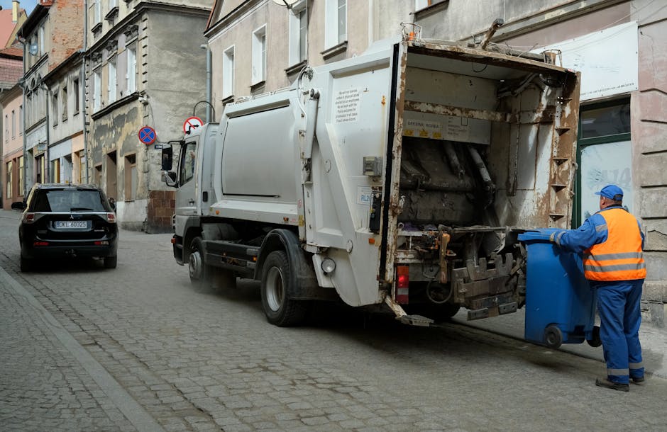 Three large black garbage bags and a rectangular cardboard box, all filled with waste materials, are situated on the edge of a paved street or sidewalk area. The bags appear to be made of thick, crink